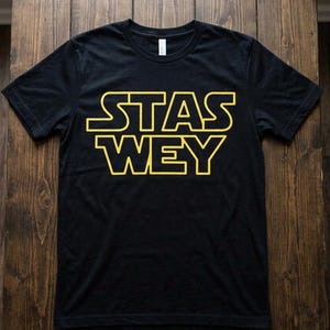Può includere: T-shirt girocollo nera con le parole "STAS WEY" in testo giallo stilizzato. Il testo è delineato in giallo. La maglietta è realizzata in un materiale morbido.