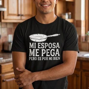 Op de afbeelding: Zwart t-shirt met de tekst "MI ESPOSA ME PEGA PERO ES POR MI BIEN" en een afbeelding van een koekenpan. De persoon die het shirt draagt, bevindt zich in een keuken.
