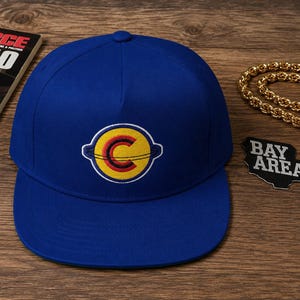 May include: A royal blue baseball cap with a flat brim. The cap features a circular patch with a yellow background, a red and yellow "C" design, and a white outline. The cap is a classic style.