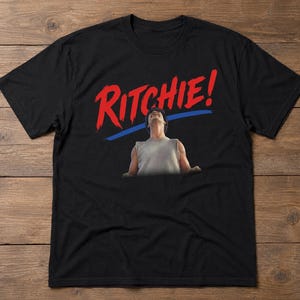 May include: Black t-shirt featuring the word "Ritchie!" in red, stylized lettering above an image of a person looking upwards. A blue line underlines the text. The person is wearing a gray tank top.