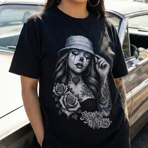 K&ouml;nnte beinhalten: Schwarzes T-Shirt mit einer Graustufen-Grafik einer Frau mit Clown-Make-up, Hut und T&auml;towierungen. Das Design beinhaltet Rosen und den Text "Loyal to the Game". Das T-Shirt hat kurze &Auml;rmel.