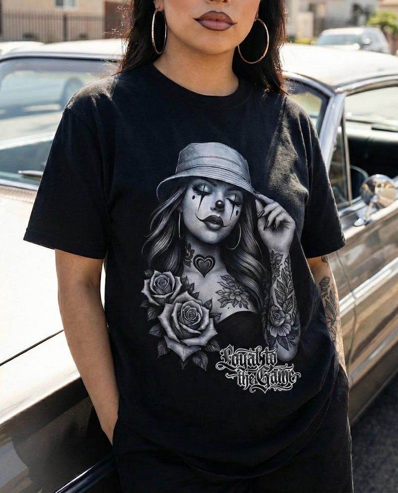 K&ouml;nnte beinhalten: Schwarzes T-Shirt mit einer Graustufen-Grafik einer Frau mit Clown-Make-up, Hut und T&auml;towierungen. Das Design beinhaltet Rosen und den Text "Loyal to the Game". Das T-Shirt hat kurze &Auml;rmel.