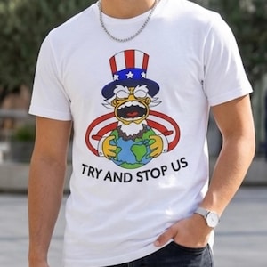 Könnte beinhalten: Weißes T-Shirt mit einem Cartoon-Design von Uncle Sam, der die Erde hält und einen wütenden Gesichtsausdruck zeigt. Die Grafik verwendet Rot-, Weiß- und Blautöne, mit dem Text "TRY AND STOP US" darunter.