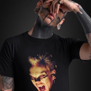 May include: Black t-shirt with a close-up of a vampire's face with fangs bared and a menacing expression. The person wearing the shirt has skull face tattoos and dark tattoos on their arms. The background is a dark grey.