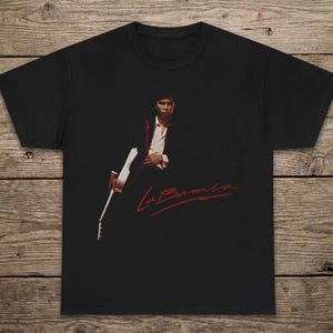 May include: Black t-shirt featuring a musician holding a guitar. The image on the shirt shows a man in a suit and white shirt. The text "La Bamba" is written in red script across the bottom of the image.