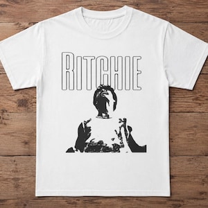 May include: White t-shirt with the name "RITCHIE" in large, outlined black letters across the top. Below the name is a black and white graphic of a person with their hands up. The shirt is made of a soft material.