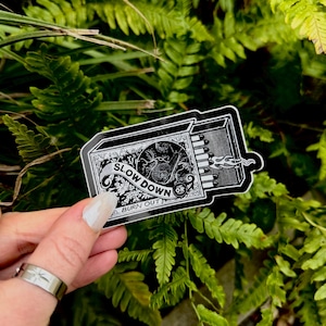 Op de afbeelding: Zwart-witte sticker met een lucifersdoosontwerp met de tekst "SLOW DOWN OR BURN OUT". De sticker heeft een gedetailleerde illustratie van een slak en bloemelementen. De lucifers zijn aangestoken, met vlammen die uit de doos komen.