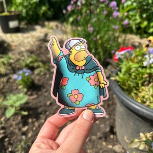 May include: A yellow cartoon character wearing a blue and pink floral dress, a black cape, and a white hat. The character is holding up one finger and has a surprised expression on their face.