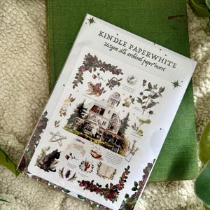 Peut inclure: Un livre vert &agrave; couverture rigide avec un insert d&eacute;coratif illustrant une maison, un chat noir et des &eacute;l&eacute;ments botaniques. L'insert porte la mention "KINDLE PAPERWHITE 265gsm silk archival paper insert."