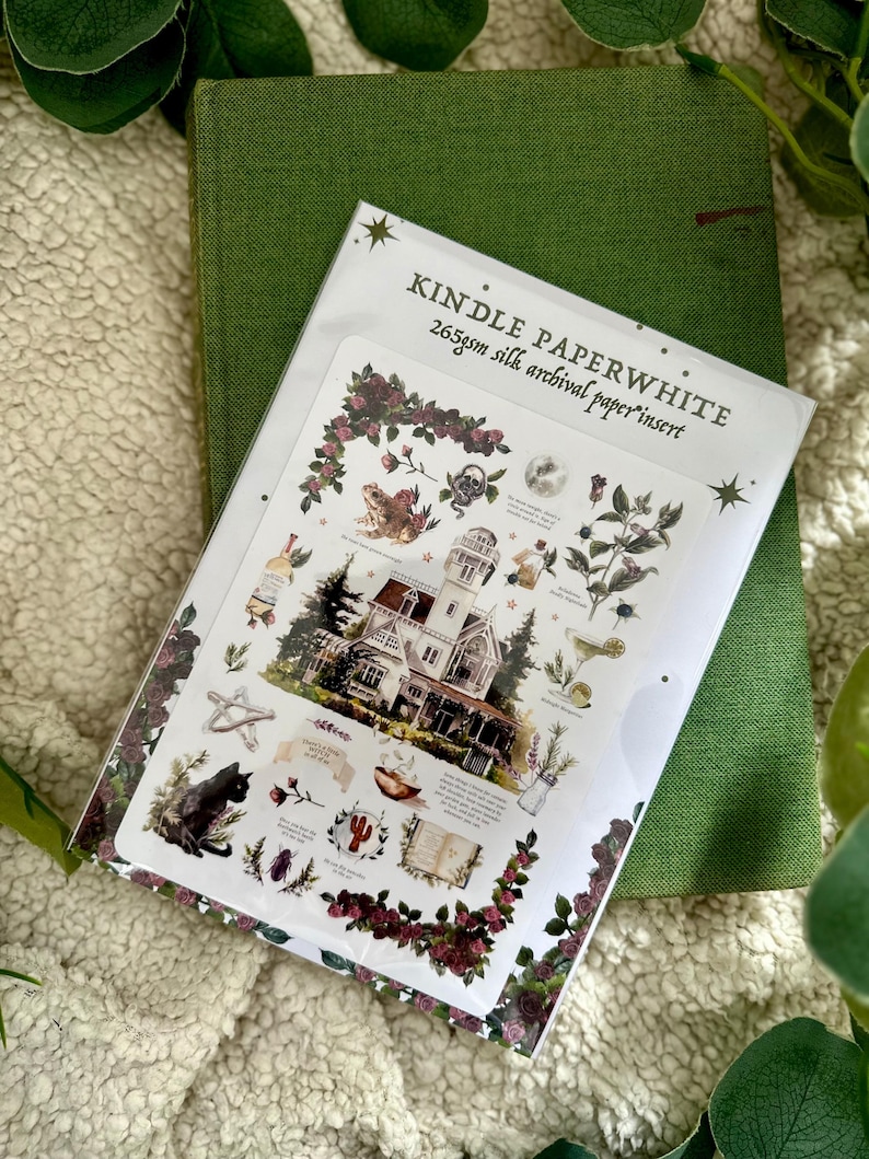 Peut inclure: Un livre vert &agrave; couverture rigide avec un insert d&eacute;coratif illustrant une maison, un chat noir et des &eacute;l&eacute;ments botaniques. L'insert porte la mention "KINDLE PAPERWHITE 265gsm silk archival paper insert."