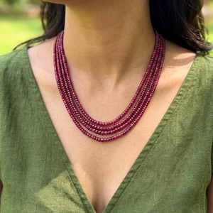 Op de afbeelding: Een meerlagige ketting bestaande uit kleine, ronde, dieprode kralen. De ketting wordt gedragen over een groene top, wat een contrast creëert. De kralen lijken gelijkmatig van grootte en dicht op elkaar geregen.