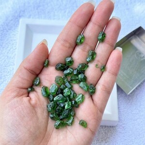 Può includere: Una manciata di pietre preziose verdi grezze e non tagliate. Le gemme sono piccole, di forma irregolare e di un colore verde smeraldo intenso. Sono tenute in una mano aperta, con un vassoio bianco e un piccolo contenitore di metallo sullo sfondo.