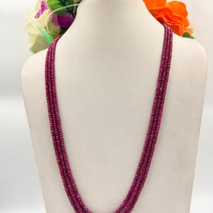 Op de afbeelding: Een meerstrengs ketting bestaande uit kleine, ronde, dieprode kralen. De ketting wordt getoond op een witte mannequin buste, met kleurrijke bloemen op de achtergrond. De kralen hebben een consistente grootte en vorm.