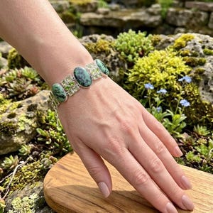 May include: A silver bracelet with oval and round green gemstones. The bracelet has a delicate, openwork design and is displayed on a wooden surface. The gemstones are a mix of dark and light green hues.