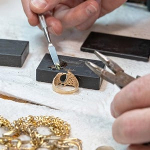 May include: A goldsmith working on a gold ring with a dark gemstone. The ring is being worked on with a small tool, and a gold chain is visible in the foreground. The ring has an oval-shaped gemstone and a detailed design.