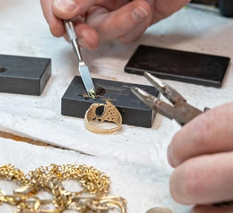 May include: A goldsmith working on a gold ring with a dark gemstone. The ring is being worked on with a small tool, and a gold chain is visible in the foreground. The ring has an oval-shaped gemstone and a detailed design.