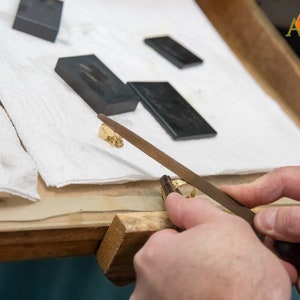 May include: A goldsmith uses a file to shape a small gold ring. The workspace includes gold jewelry, a ring, and several dark rectangular blocks on a white cloth. The logo "A2AR" is visible in the top right corner.