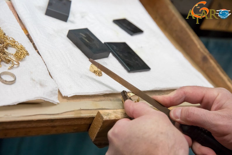 May include: A goldsmith uses a file to shape a small gold ring. The workspace includes gold jewelry, a ring, and several dark rectangular blocks on a white cloth. The logo "A2AR" is visible in the top right corner.