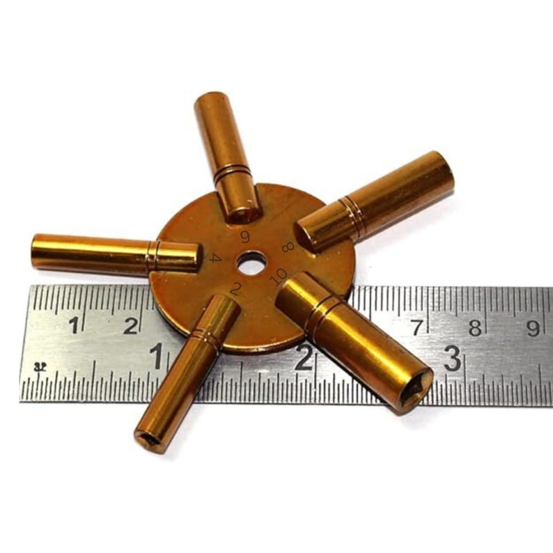 May include: A gold-toned watch key winding tool with five different sized prongs. The prongs are numbered 2, 4, 8, 9, and 10. The tool is measured against a ruler.