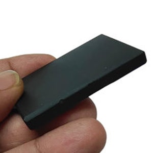 May include: A small, rectangular black object with a smooth surface. The object is held in a person's hand against a white background. The object appears to be made of a solid material.