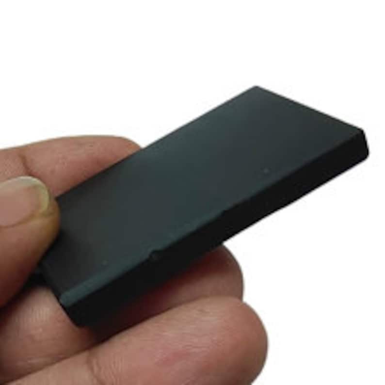 May include: A small, rectangular black object with a smooth surface. The object is held in a person's hand against a white background. The object appears to be made of a solid material.