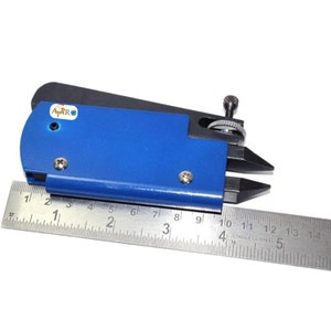 May include: A blue and black metal tool with a small knob on the side. The tool is measured against a ruler that is marked in inches. The tool is labelled "AARO".