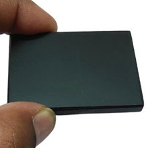 May include: A rectangular, black, polished stone held between fingers. The stone has a smooth, reflective surface. The edges of the stone are slightly beveled. The stone is approximately 3 inches long and 2 inches wide.