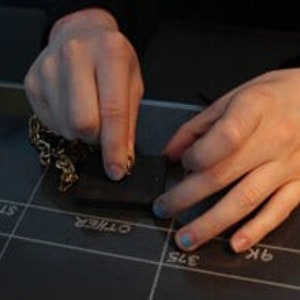 May include: A person's hands working with a small, dark rectangular object, possibly jewelry, on a grid-lined surface. A chain is visible on the left. The surface has text and markings.