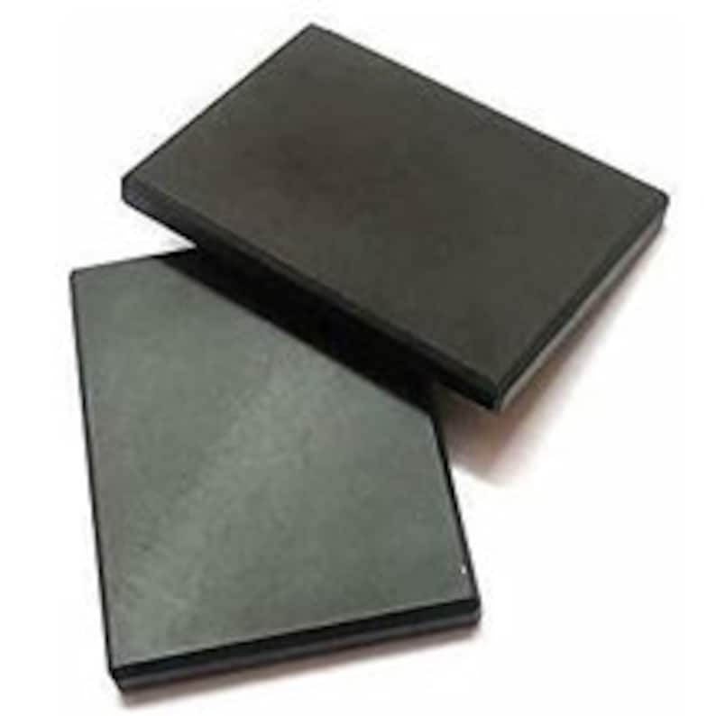 May include: Two rectangular, black soapstone coasters are stacked on a white background. The coasters have a smooth, matte finish and a slightly beveled edge. They appear to be new and unused.