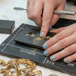 May include: A goldsmith's hands working on a gold chain. The hands are using tools to manipulate the chain on a black surface. The surface has markings and the text "OTHER" and "375". Gold dust is scattered around the chain.