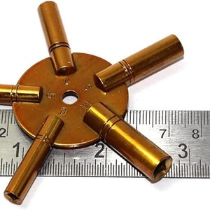 May include: A gold-toned watch key winding tool with five different sized prongs. The tool is measured against a ruler with markings in inches.