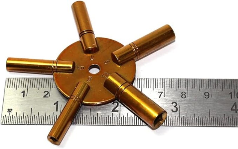 May include: A gold-toned watch key winding tool with five different sized prongs. The tool is measured against a ruler with markings in inches.