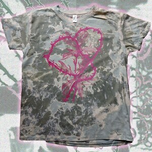 May include: A tie-dye t-shirt with a pink heart-shaped design. The heart is formed by a thorny vine and contains a floral element. The shirt has a crew neck and short sleeves. The background has a mottled, faded appearance.