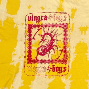 Peut inclure: T-shirt jaune tie-dye avec un motif rouge et noir. Le motif comprend le texte "viagra boys" en police gothique et une créature ressemblant à un insecte avec des pointes. Le t-shirt a un col rond et des manches courtes.