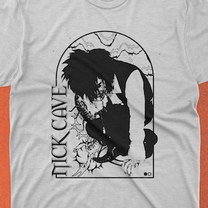 May include: A gray t-shirt with a black and white graphic print of a person with long hair. The graphic is in an arched frame with the text "NICK CAVE" printed vertically on the left side.