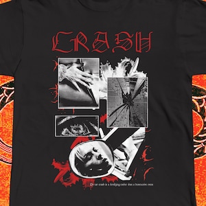 Op de afbeelding: Een zwart t-shirt met korte mouwen en een rode grafische print met het woord CRASH in een gestileerd lettertype. Het ontwerp bevat vier zwart-wit fotopanelen met een hand, een verbrijzeld oppervlak, een oog en een gezicht in een spiegel.
