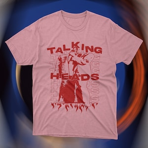 Vintage-inspired pink Talking Heads t-shirt featuring bold red graphic print of David Byrne in oversized suit with flames and repeating text design. Retro new wave band tee, unisex fit