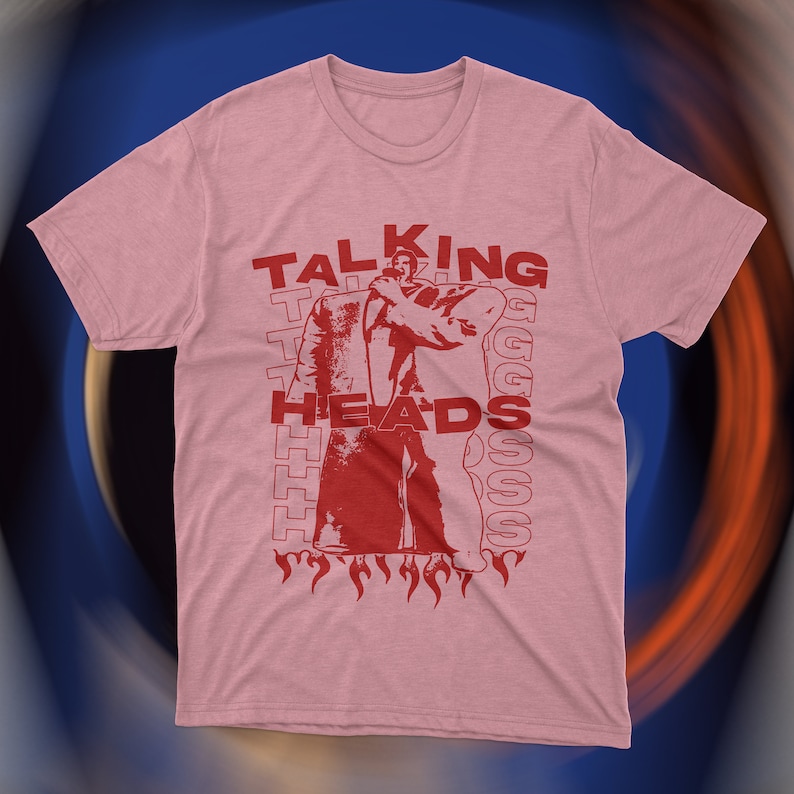 Vintage-inspired pink Talking Heads t-shirt featuring bold red graphic print of David Byrne in oversized suit with flames and repeating text design. Retro new wave band tee, unisex fit