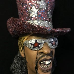 May include: A sculpture of a person wearing a red glitter top hat with silver stars and a silver band. The person is also wearing silver sunglasses with red star accents. The sculpture has a beard and is smiling with teeth showing.