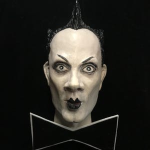 May include: A sculpted bust of a pale-skinned figure with a black mohawk hairstyle, dark lipstick, and a white bow tie. The figure has a stern expression with dark eyebrows and wide eyes. The background is solid black.
