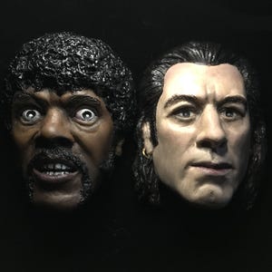 May include: Two detailed busts of male figures. One has dark skin, a mustache, and curly hair, with wide eyes and an open mouth. The other has light skin, long hair, and a serious expression, with a gold earring.
