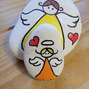 Puede incluir: Dos piedras blancas pintadas con diseños de ángeles. La piedra más grande tiene un ángel amarillo con un halo y alas. La piedra más pequeña tiene un ángel naranja con un halo y alas. Ambos ángeles tienen corazones rojos a su alrededor.