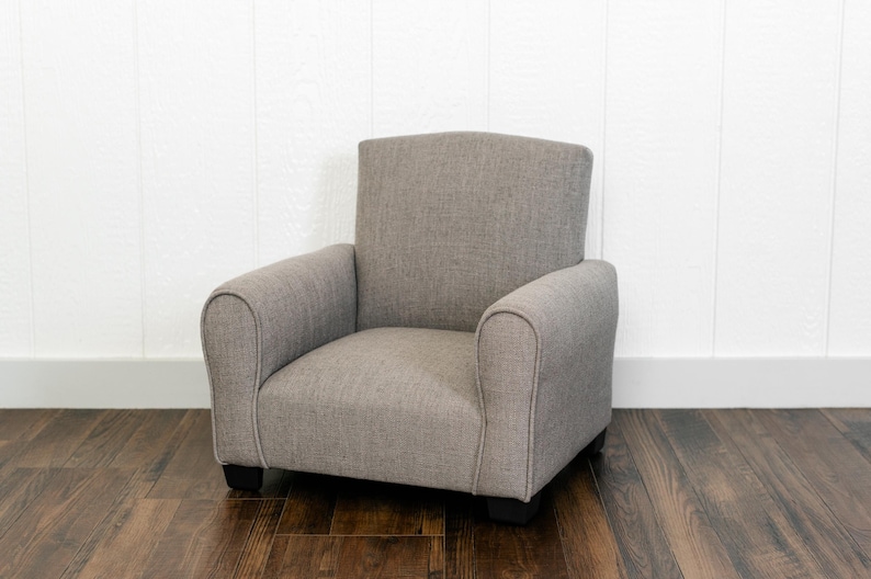 May include: A small, upholstered armchair featuring a textured, light gray fabric finish. The chair has a high, straight backrest, rounded armrests, and a low-profile seat cushion, supported by four short, dark-colored feet at the base.