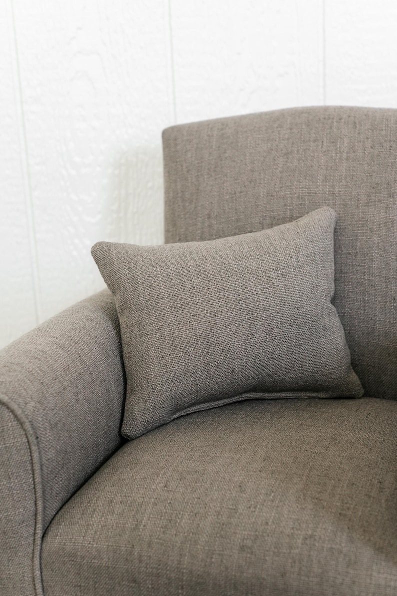 May include: A rectangular throw pillow made of a textured, taupe-colored fabric. The pillow features a simple, solid-colored design with visible woven threads, resting against the matching upholstered back and seat of a chair.
