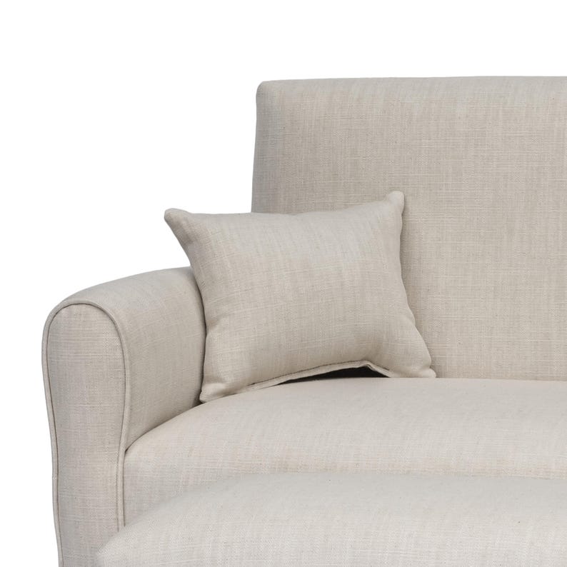May include: A beige sofa with a textured fabric finish and a matching square throw pillow resting against the armrest. The sofa features a rounded arm design and a clean, neutral appearance with a consistent woven pattern across all surfaces.
