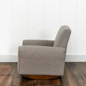 May include: A side view of a gray upholstered rocking chair with a textured fabric finish. The chair features a high backrest, straight armrests, and a curved wooden base designed for a smooth rocking motion, presented in a neutral color palette.