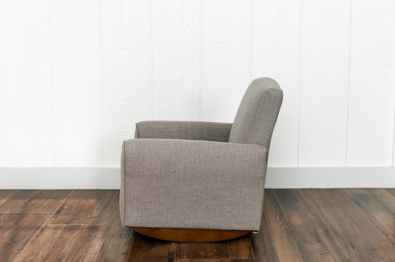 May include: A side view of a gray upholstered rocking chair with a textured fabric finish. The chair features a high backrest, straight armrests, and a curved wooden base designed for a smooth rocking motion, presented in a neutral color palette.