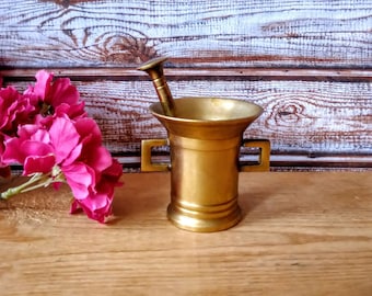 May include: A brass mortar and pestle with a wooden base. The mortar is cylindrical in shape with a wide base and a narrow top. The pestle is a long, thin rod with a rounded end. Both the mortar and pestle are shiny and have a slightly aged appearance.