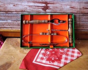 Op de afbeelding: Een set van drie keukenhulpmiddelen in een groene en oranje doos. De hulpmiddelen omvatten een flesopener, een kurkentrekker en een tang. De hulpmiddelen hebben houten handvatten.