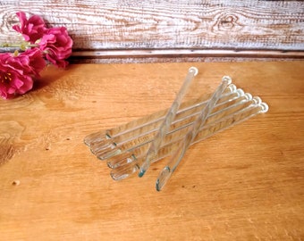 May include: A set of eight clear glass stirring rods, arranged in a crisscross pattern on a wooden surface.